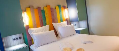 Habitación de hotel con una cama, almohadas blancas, una mesita de noche, una lámpara y un sombrero sobre la cama.