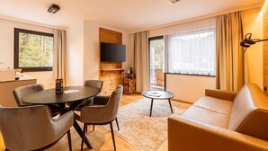 A modern living room with a round dining table, two sofas, a TV mounted on a wooden wall, and large windows with curtains. - Nice suite in Saalbach (Saalbach)