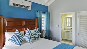 A bedroom with a wooden headboard, a bed with white linens, and blue and white patterned pillows. There is a window with blue curtains, a bedside table with a lamp, and a bathroom visible through an open door.