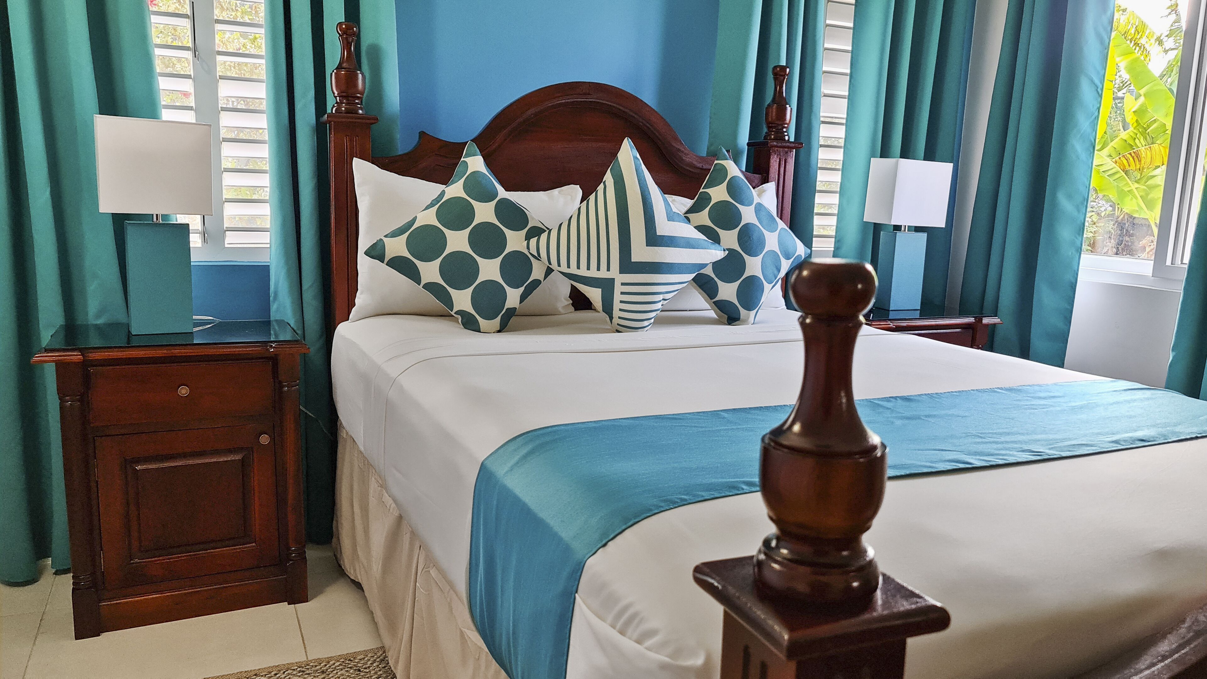 A neatly made bed with a wooden headboard, a blue bedspread, and patterned pillows. There is a nightstand with a lamp and a wooden drawer. The room has teal curtains and a view of greenery outside.