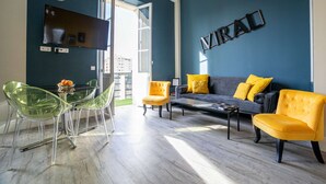 A modern living room with a blue accent wall, a sofa with yellow cushions, and a dining area with clear chairs. - Le Loft de St Jean - 2 bed and 2 bath (Cannes)