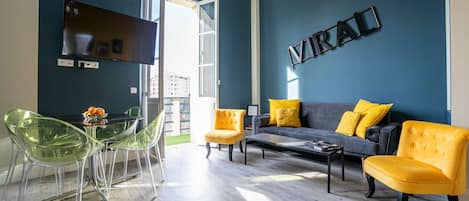 A modern living room with a blue accent wall, a sofa with yellow cushions, and a dining area with clear chairs.