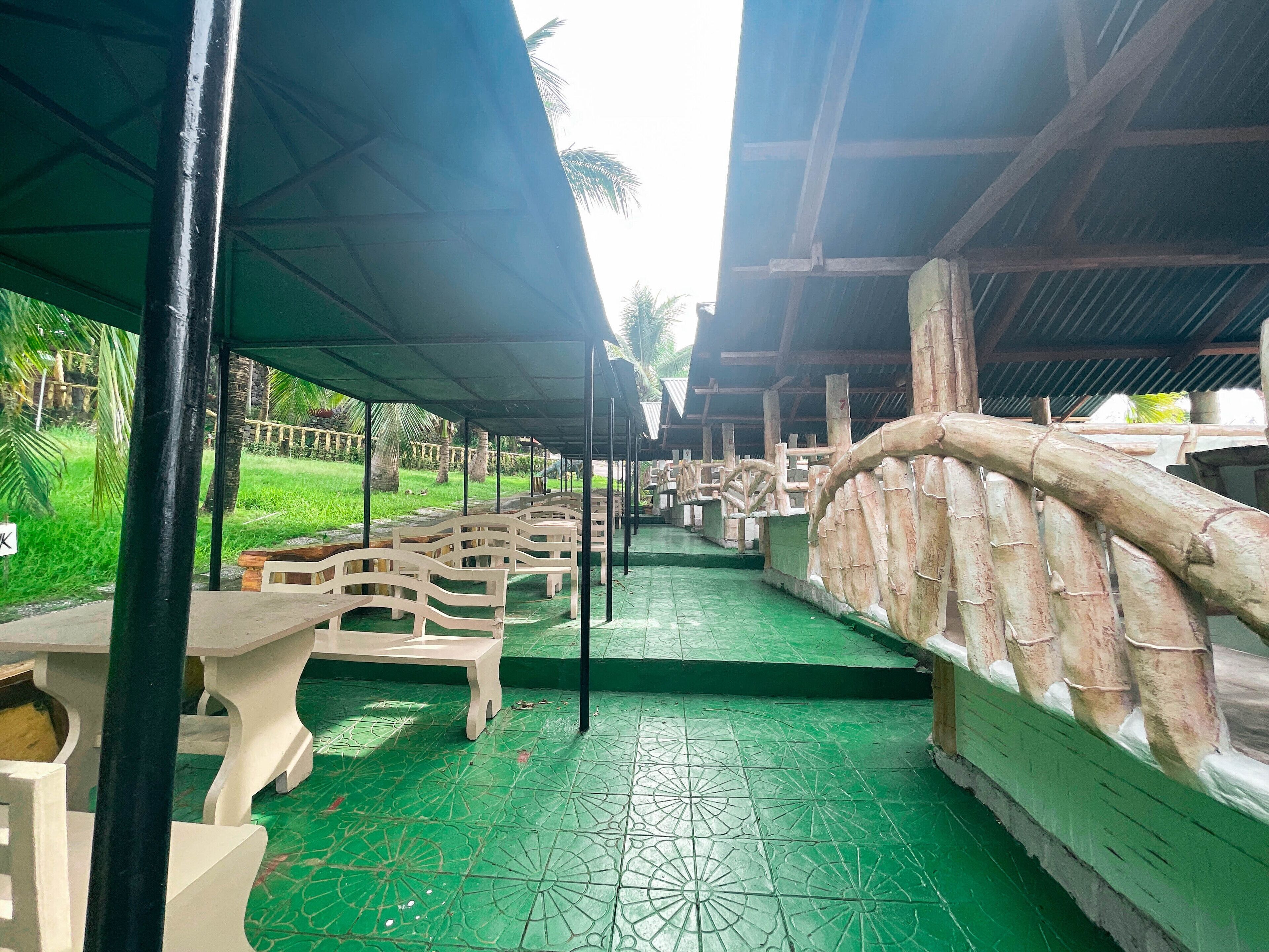A covered outdoor seating area with tables and chairs, a green tiled floor, and a bamboo railing.