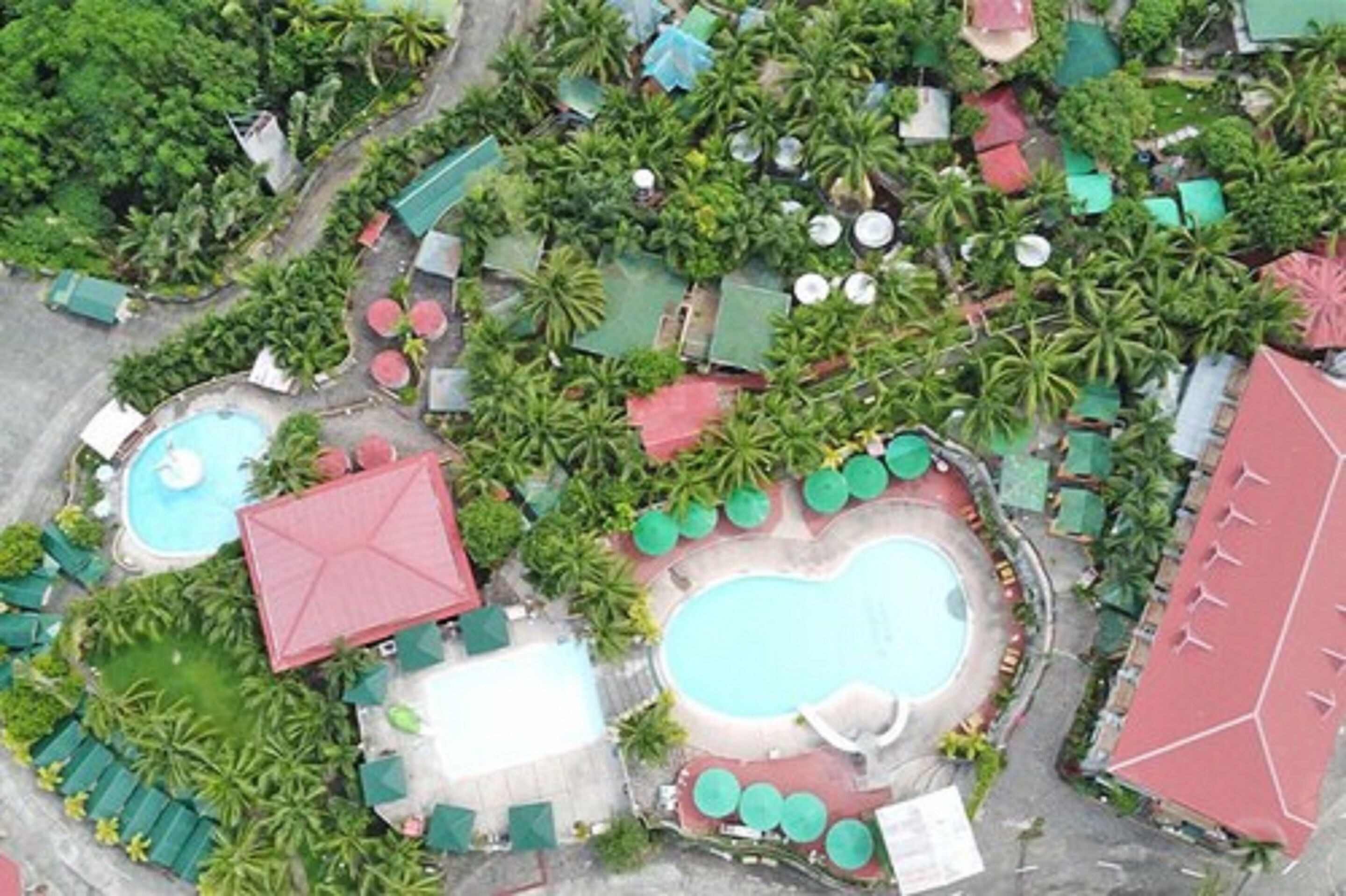 An aerial view of a resort with multiple swimming pools, green umbrellas, and palm trees.