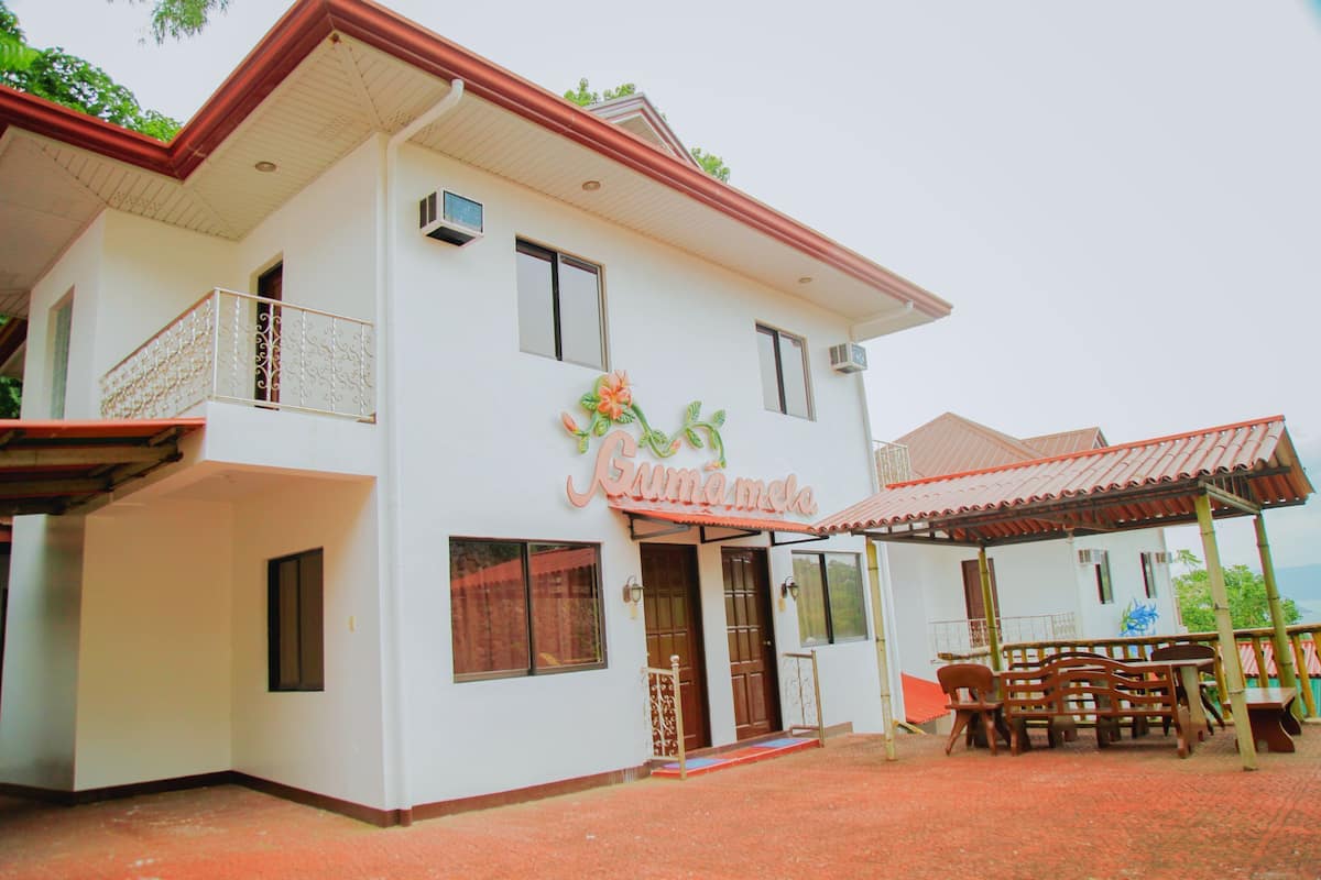A two-story building with a balcony, a covered patio with furniture, and a sign that reads "Kumamela".