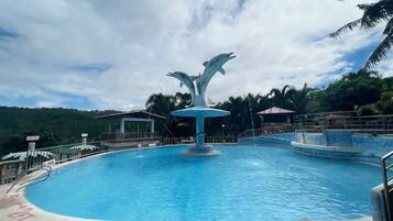 A swimming pool with a dolphin fountain, surrounded by palm trees and a view of a hilly landscape.