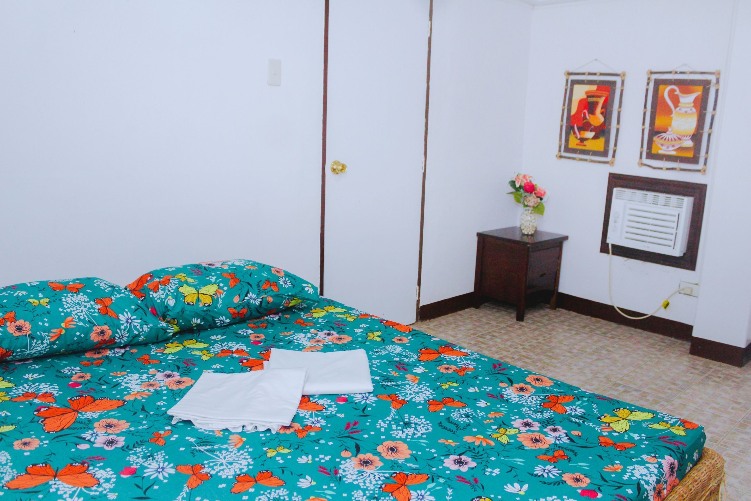 A room with a bed, a small table with a vase, and two framed pictures on the wall.
