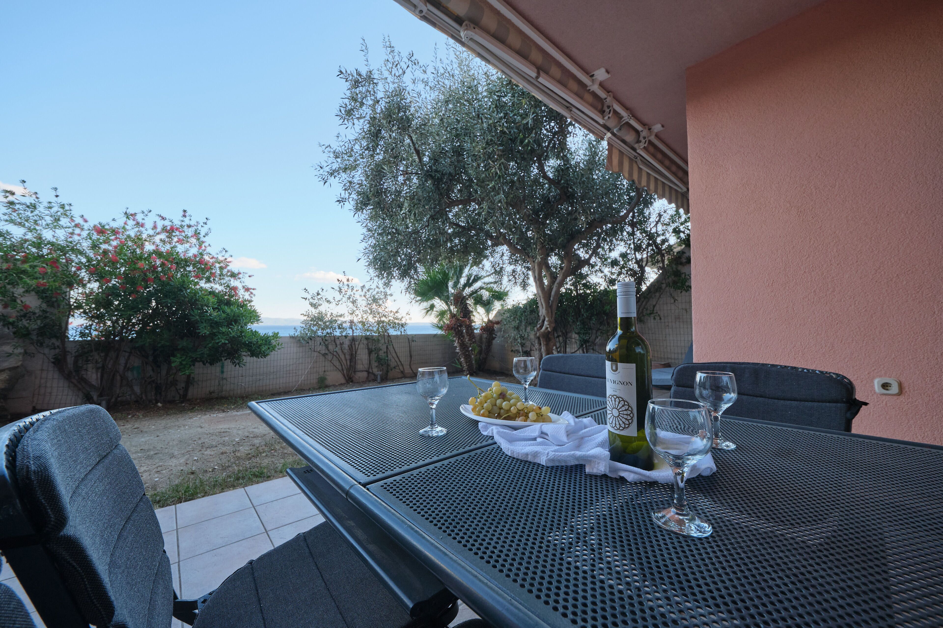 An outdoor patio with a table set for two, featuring a bottle of wine, a plate of grapes, and two glasses.