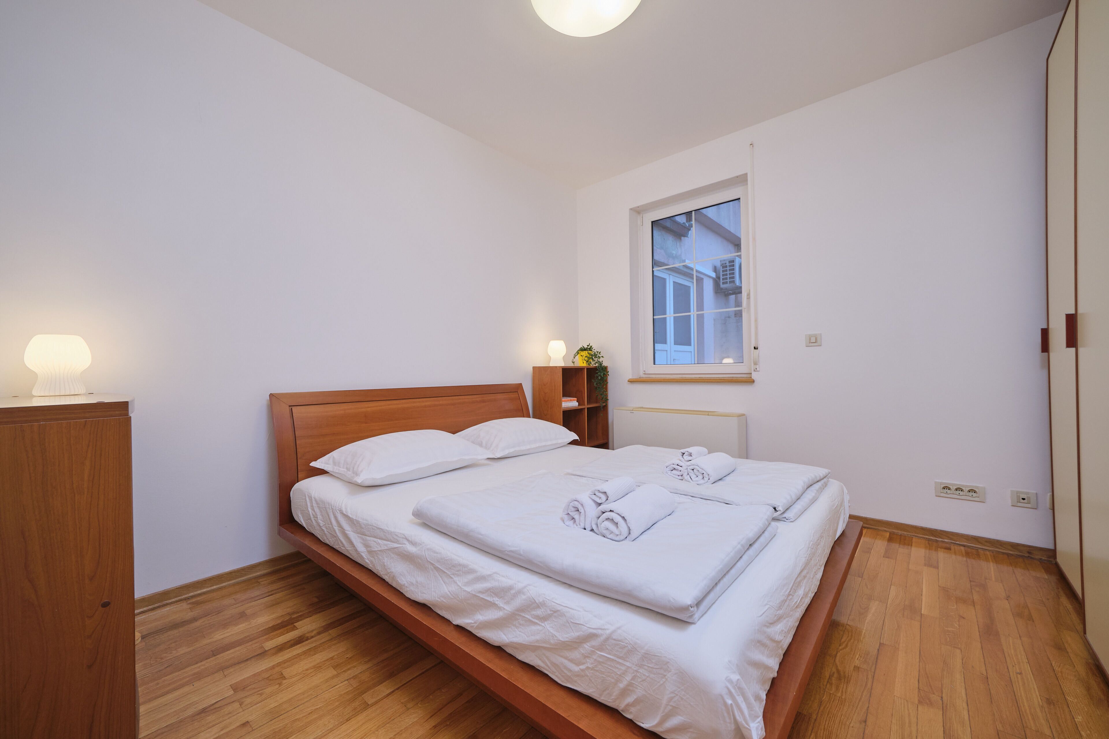 Un lit bien fait, avec du linge de lit et des serviettes blanches, une tête de lit en bois, une table de chevet avec une lampe et une fenêtre donnant sur un bâtiment à l’extérieur.