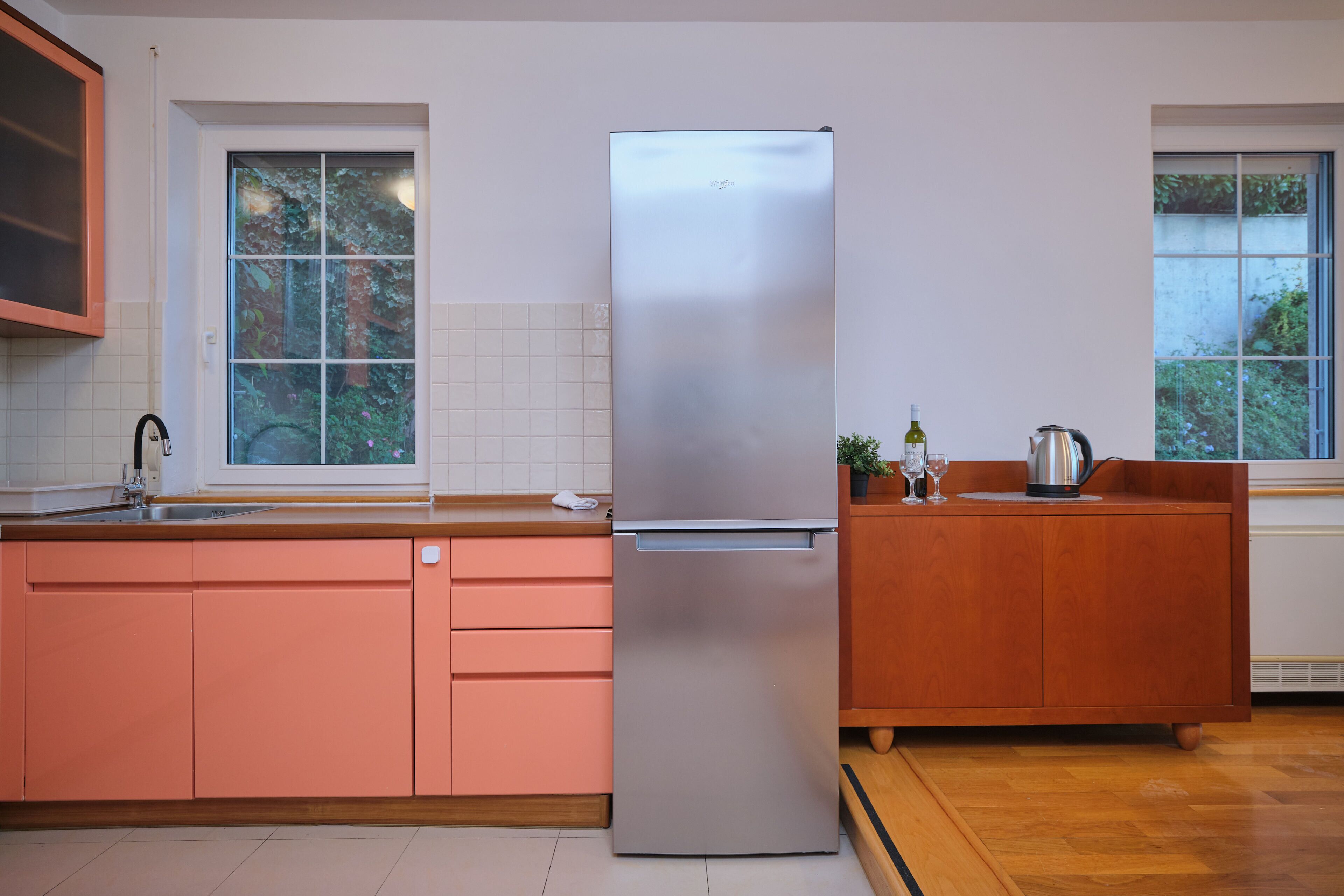 A modern kitchen with a stainless steel refrigerator, wooden cabinets, and a window with a view of greenery.