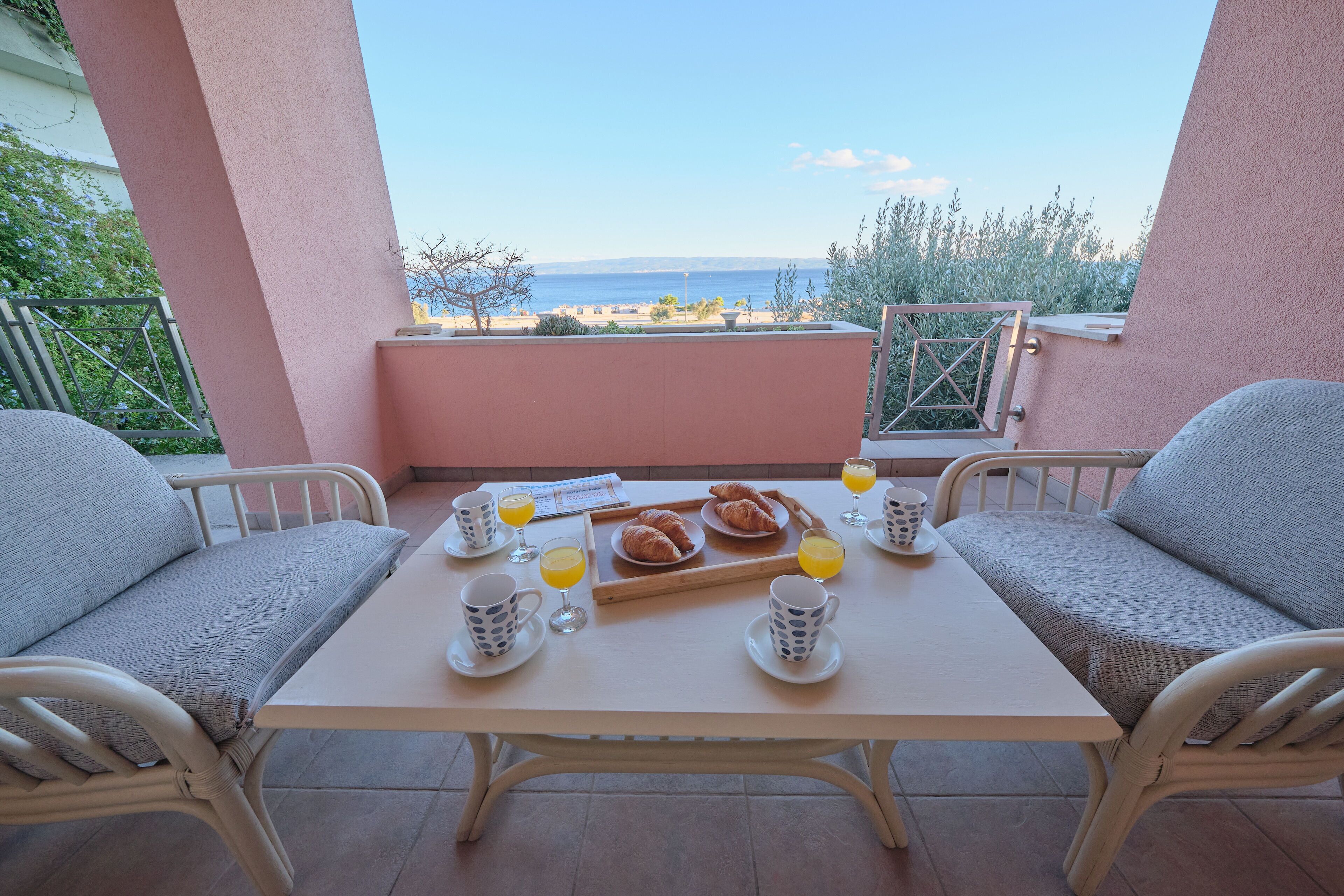 A breakfast table with croissants, orange juice, and coffee cups on a balcony overlooking the sea.