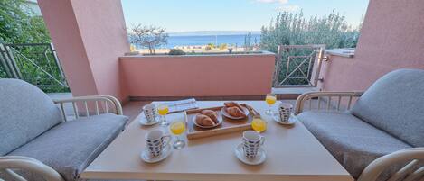 A breakfast table with croissants, orange juice, and coffee cups on a balcony overlooking the sea.