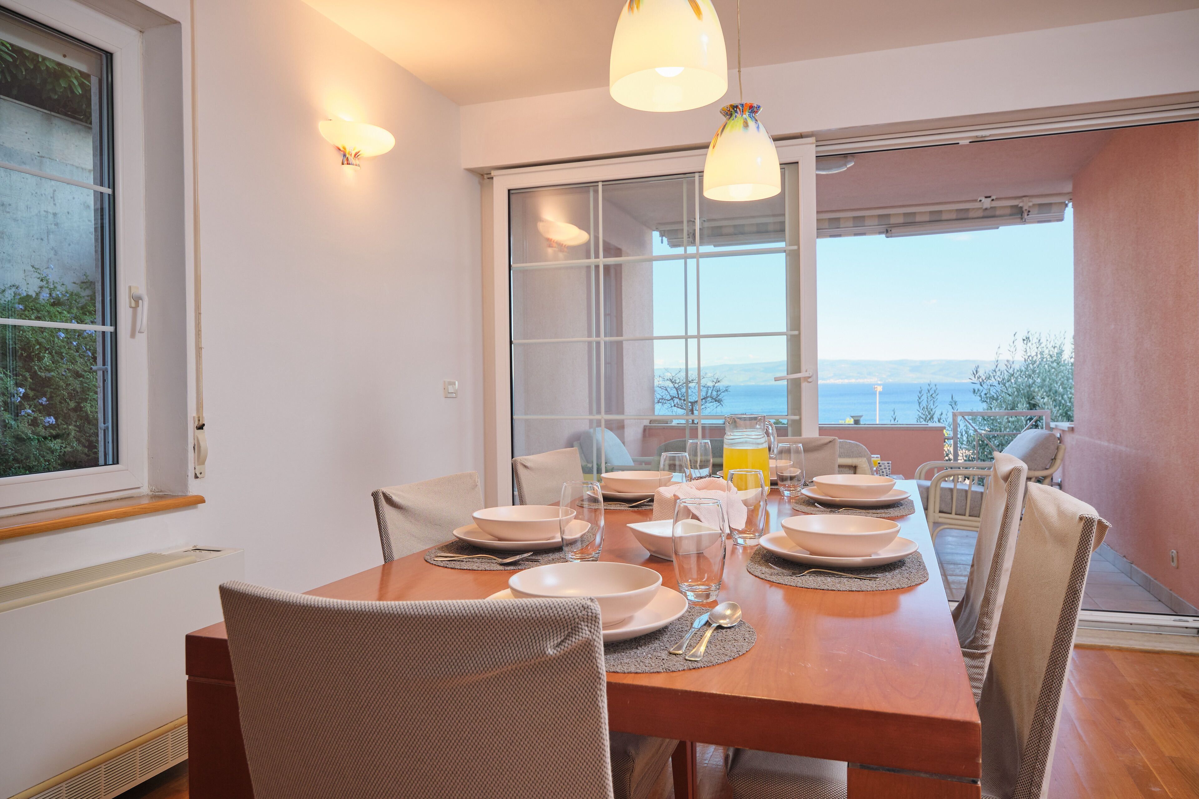 Un comedor con una mesa de madera preparada para cuatro personas, rodeado de sillas de mimbre, con vista al mar a través de amplios ventanales.