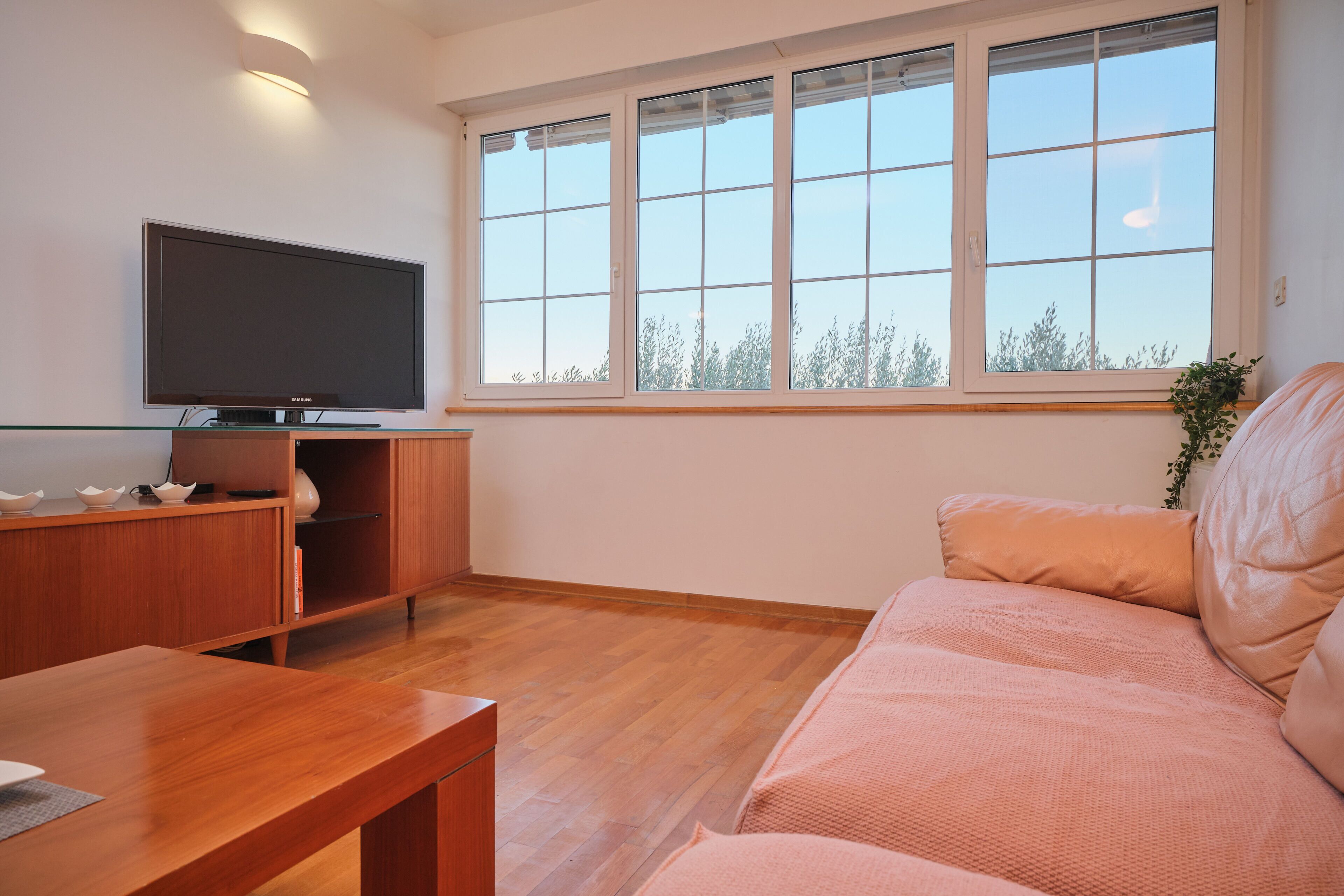 A modern living room with a flat-screen TV, wooden furniture, and a large window with a view of trees.
