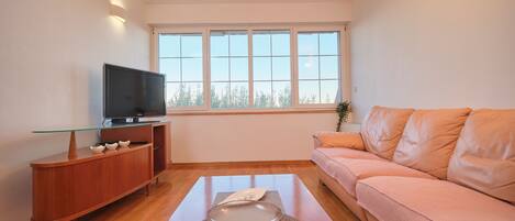 A modern living room with a flat-screen TV on a wooden cabinet, a glass-top coffee table, a pink sofa, and a large window with a view of trees.