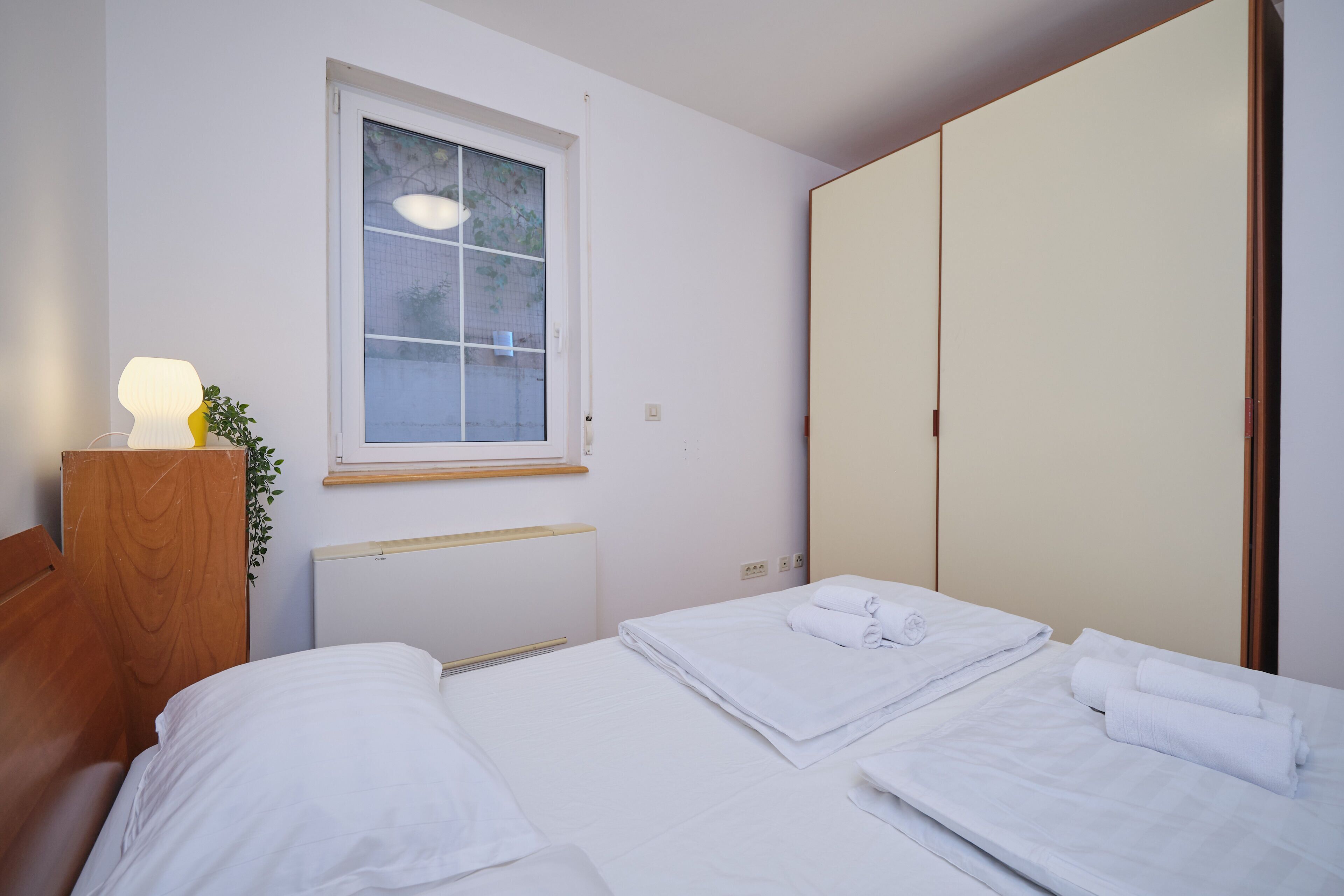 A hotel room with a bed, a bedside table with a lamp, a window, and a wardrobe.
