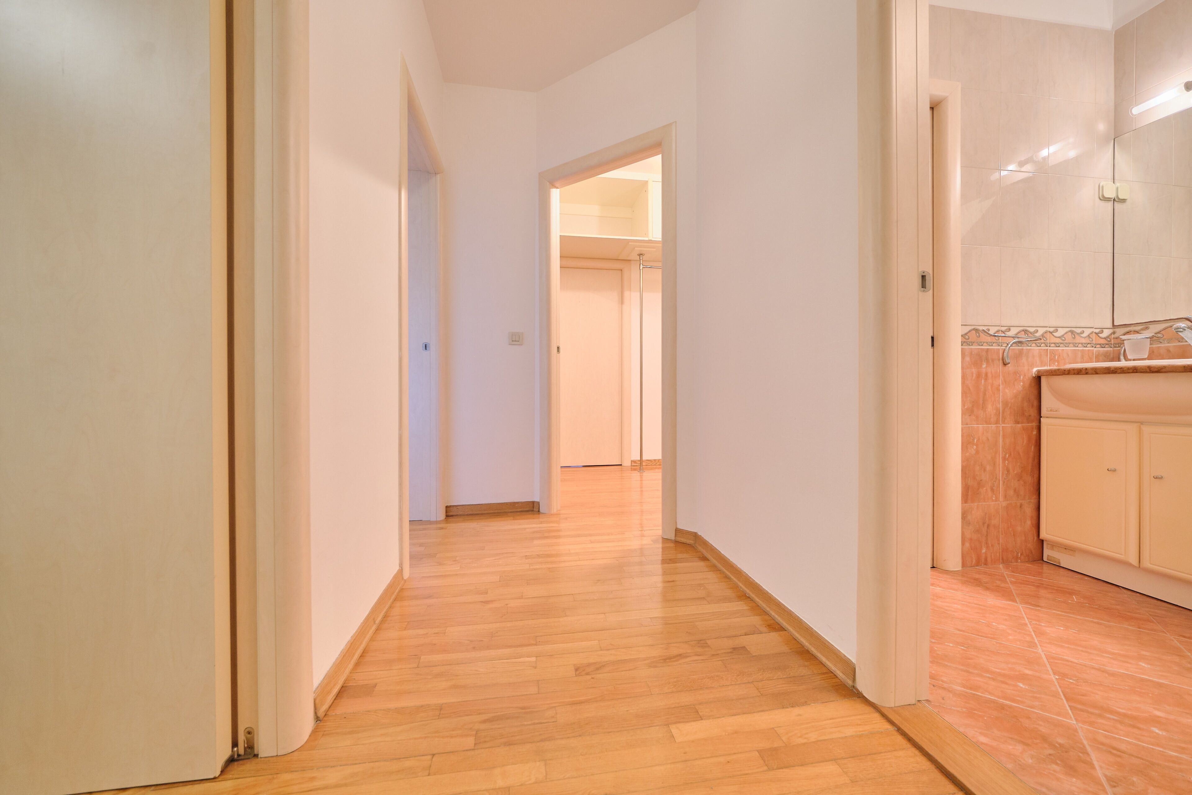 A hallway with wooden flooring leading to a bathroom with a sink and cabinet.