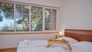 A hotel room with two beds, a wooden headboard, a window with a view of trees and the sea, and a tray with a croissant and orange juice. - Holiday Seaview Apartment by Litto (Split)