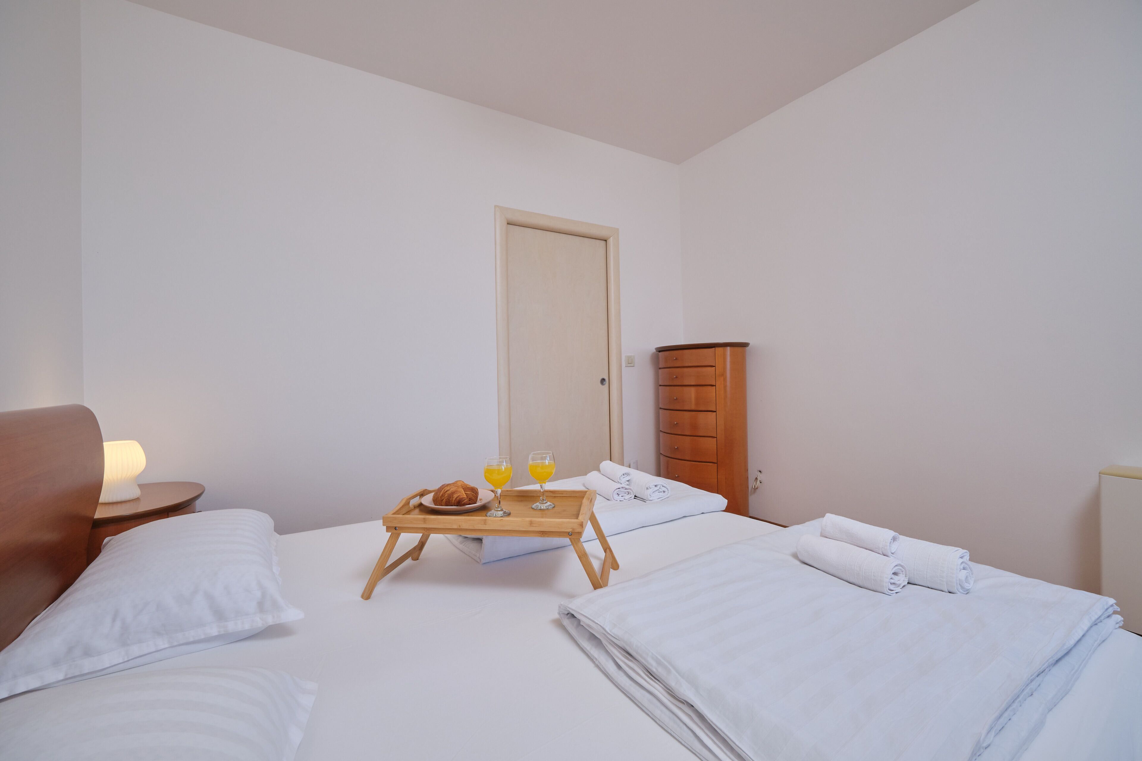 A hotel room with a bed, a wooden side table with a lamp, a wooden dresser, and a breakfast tray with a croissant, juice glasses, and a plate.