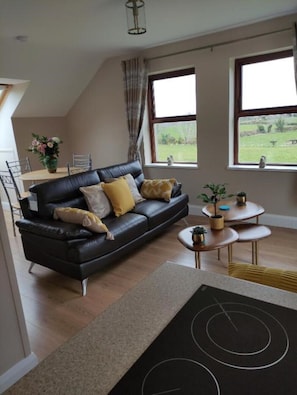 Uma sala de estar moderna com um sofá de couro preto, uma mesa de jantar e vista para um campo verde através de grandes janelas.