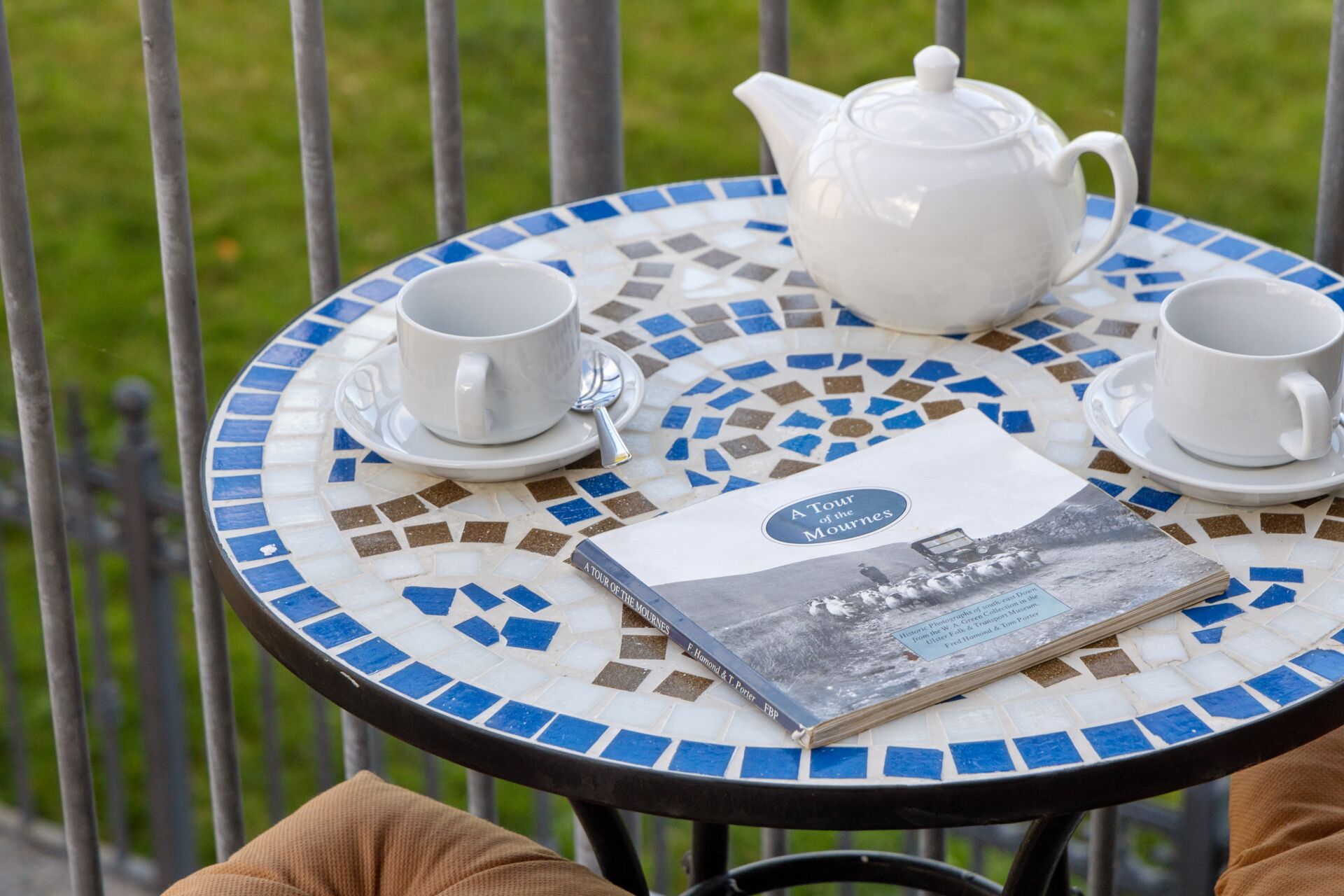 A round table with a mosaic tile design, two white cups, a white teapot, and a book titled "A Tour of the Mournes".