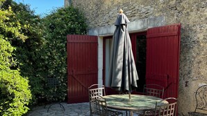 Une terrasse en pierre avec une table ronde et quatre chaises, un parasol rouge et un bâtiment aux volets rouges.