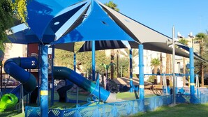 A playground with a slide and climbing structures, surrounded by a fence and palm trees. - Eilat Apartments  (Eilat)