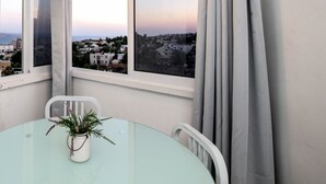 A modern room with a round glass table, a potted plant, and a view of a coastal cityscape through large windows. - Eilat Apartments  (Eilat)