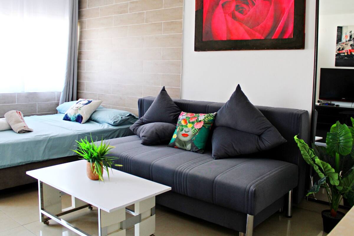 A modern living room with a grey sofa, a white coffee table, a bed with a blue bedspread, and a large red rose painting on the wall.