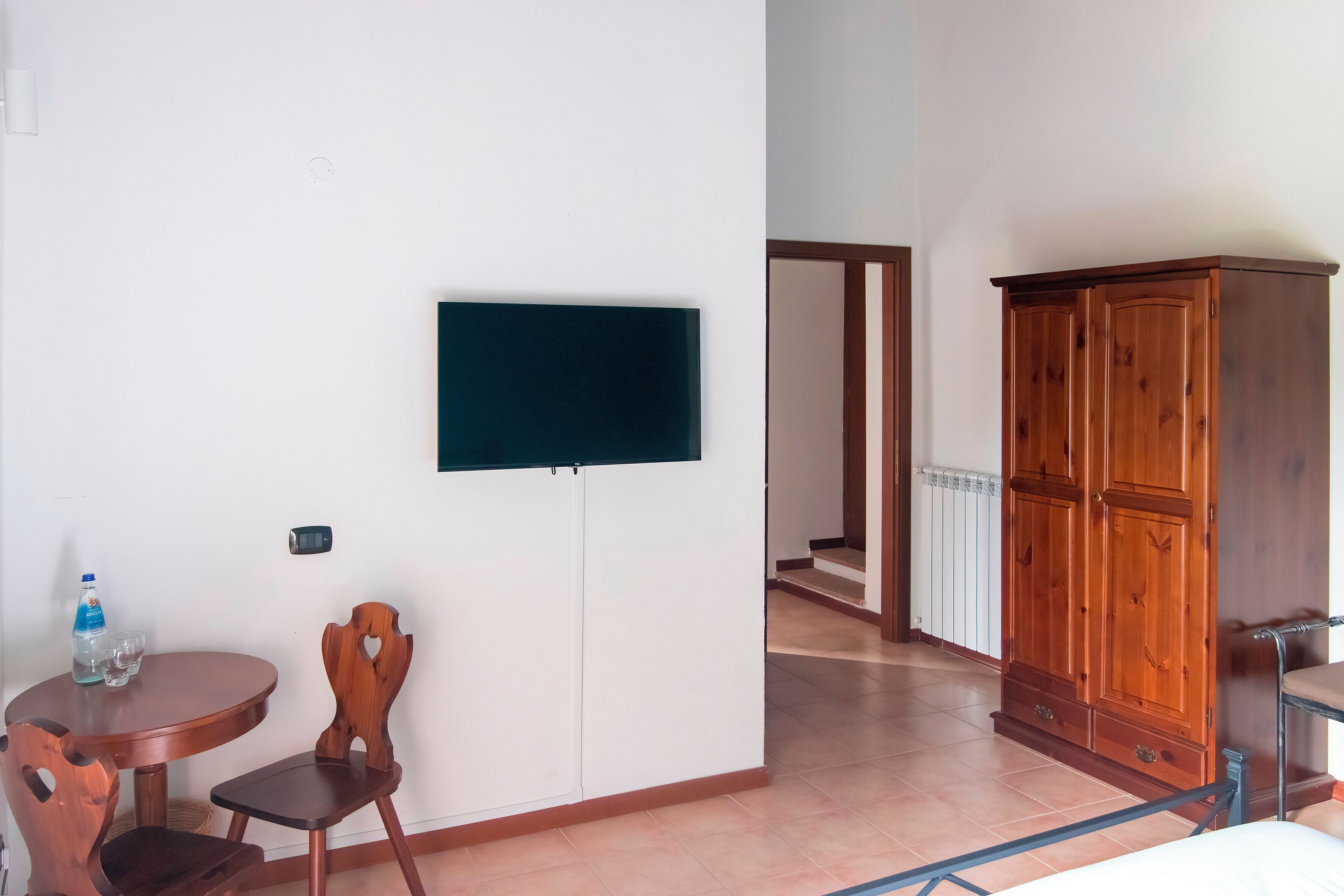 Una habitación con un armario de madera, un televisor montado en la pared, una mesa redonda de madera con dos sillas y una botella de agua sobre la mesa.
