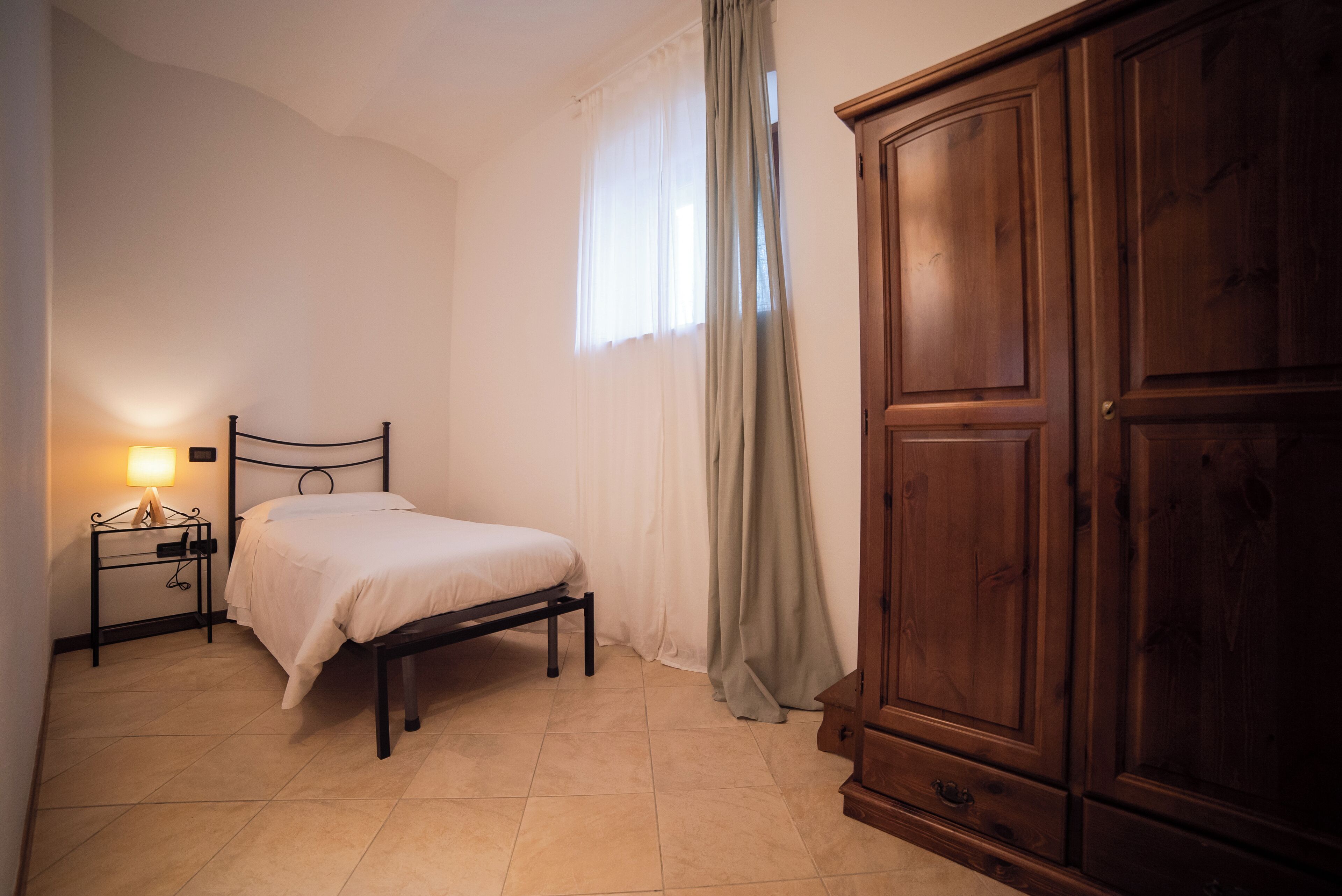 Un dormitorio con cama, mesita de noche, lámpara, ventana con cortinas y un armario de madera.