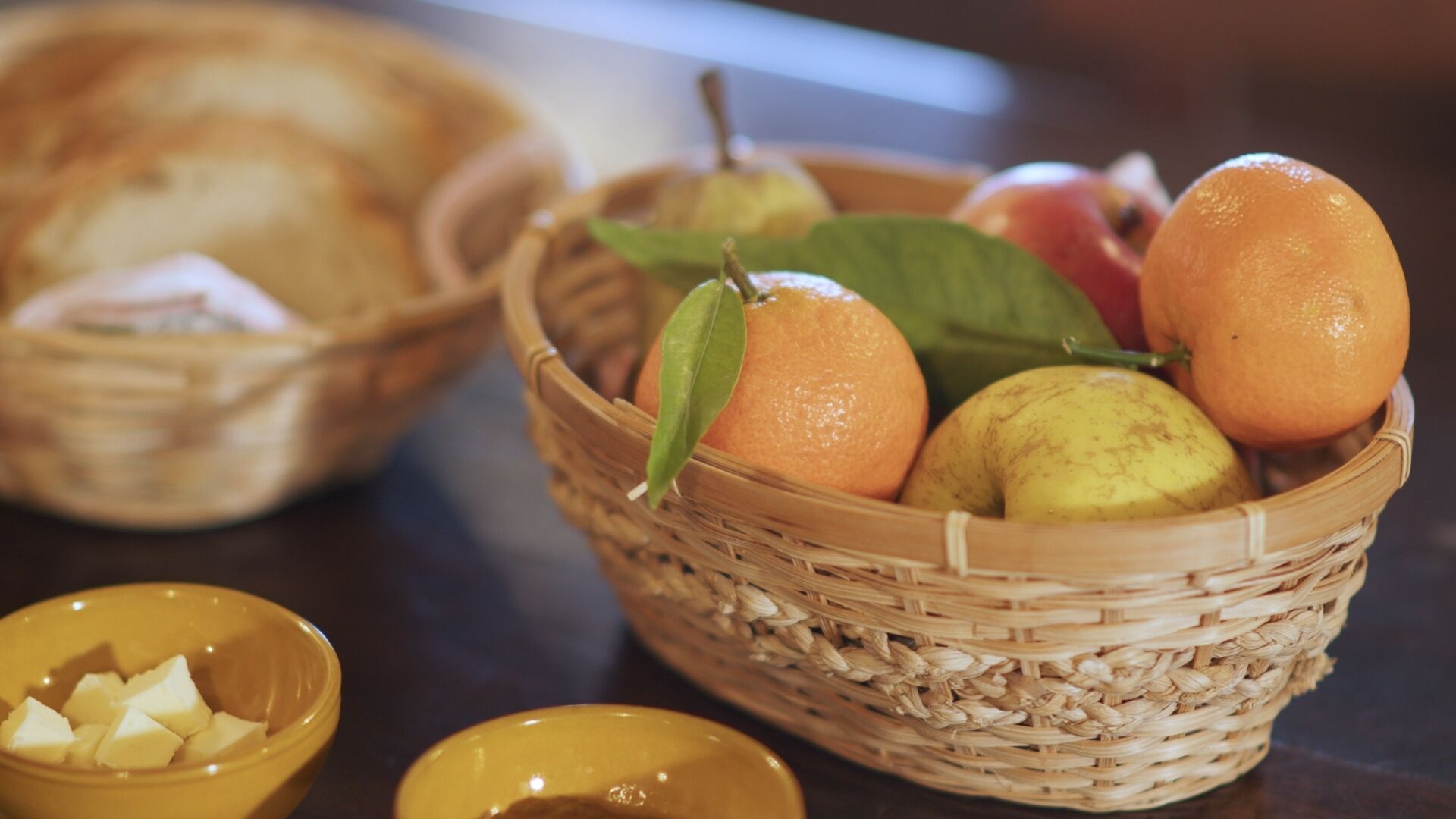 Una cesta de mimbre con naranjas, una pera y una manzana verde, junto con un bol de fruta cortada y un recipiente pequeño con mantequilla sobre una mesa.