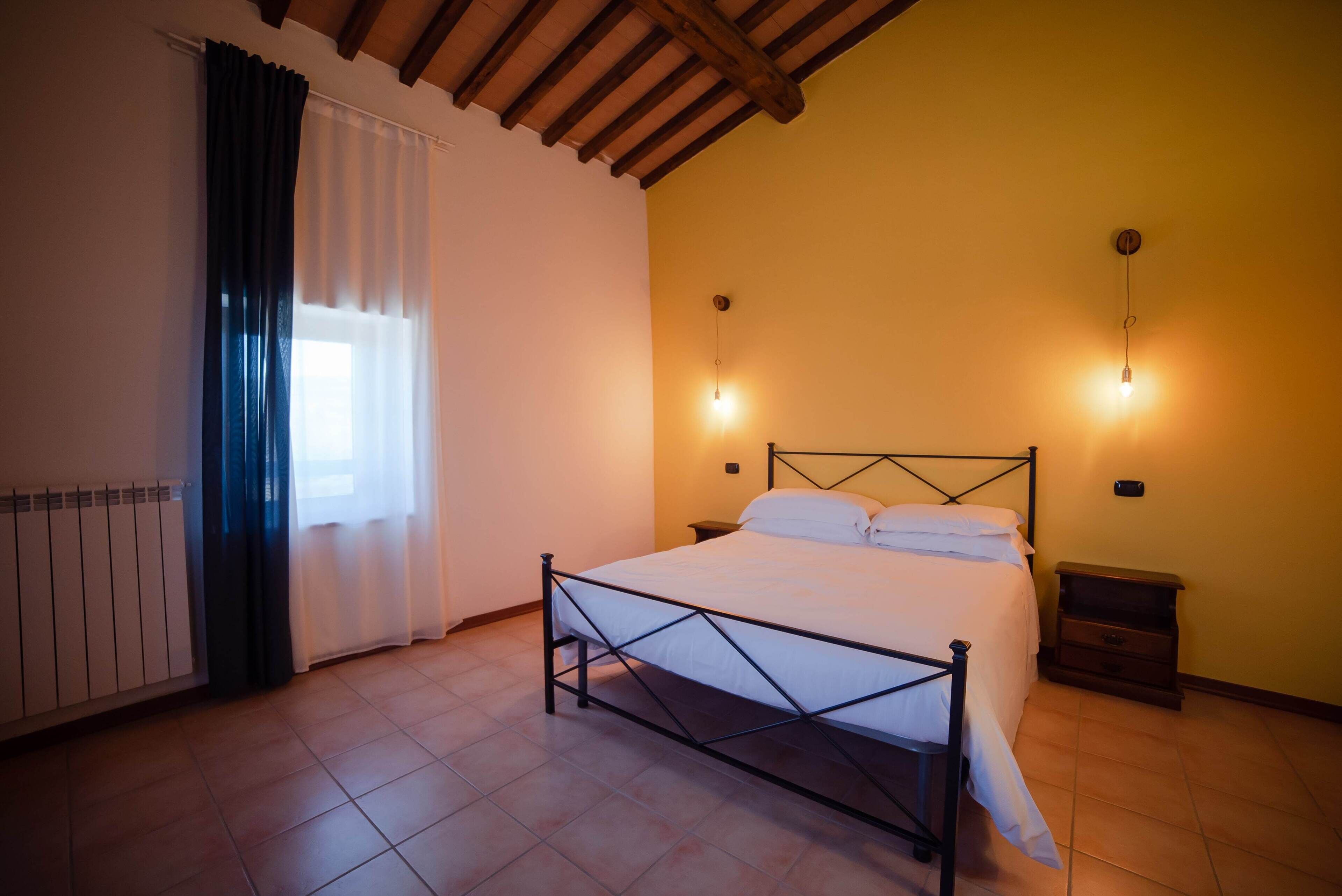 Une chambre à coucher avec un lit, une table de chevet, une lampe fixée au mur et une fenêtre avec des rideaux.