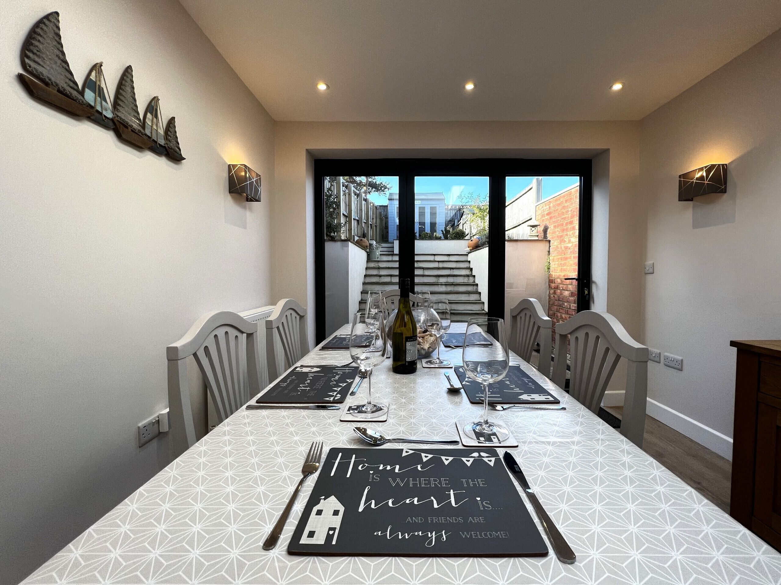 Comedor con una mesa larga dispuesta para seis personas, con un mantel blanco estampado, un posavasos negro con una cita y vista a una escalera que da al exterior.