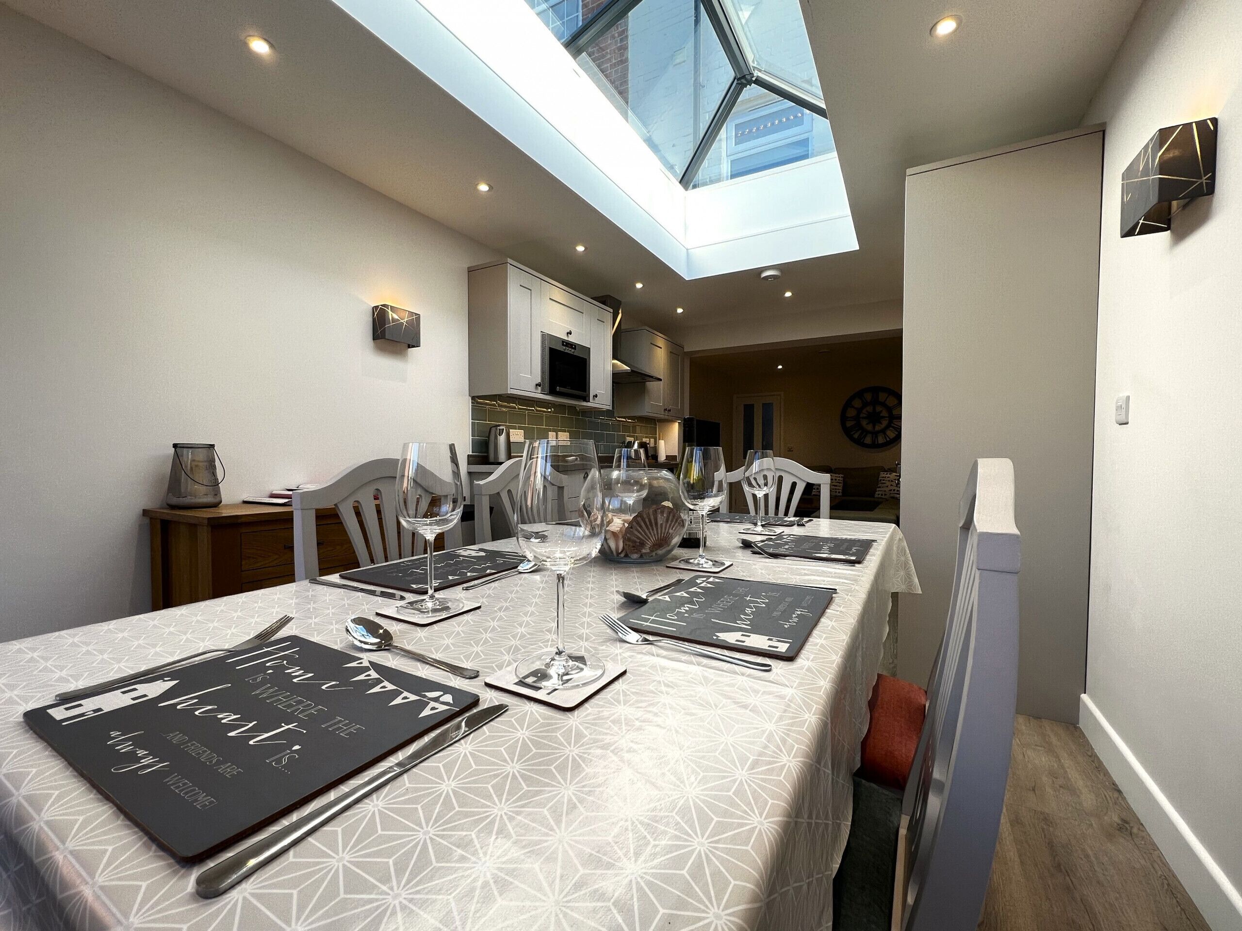 Un comedor con techo alto y tragaluz, que presenta un mantel con estampado geométrico, una mesa de comedor ordenada con cubiertos, y una vista de la cocina al fondo.