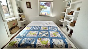 A bedroom with a bed, a window with a view, and shelves with various items. - Bouganville  (Ischia)