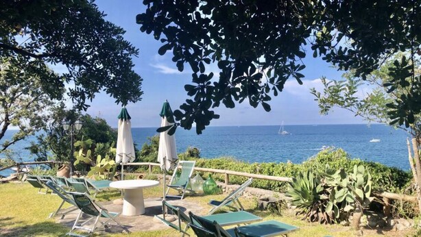 An outdoor seating area with a table and chairs, surrounded by greenery and overlooking a body of water. - Bouganville  (Ischia)