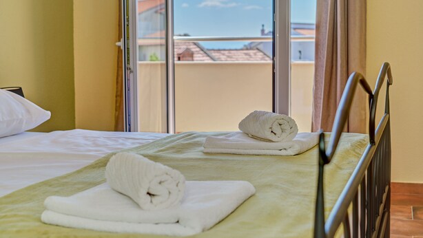 A hotel room with a bed, two rolled towels, a window with a view of buildings, and a metal bed frame. - Anovi Holiday House by Litto (Okrug)
