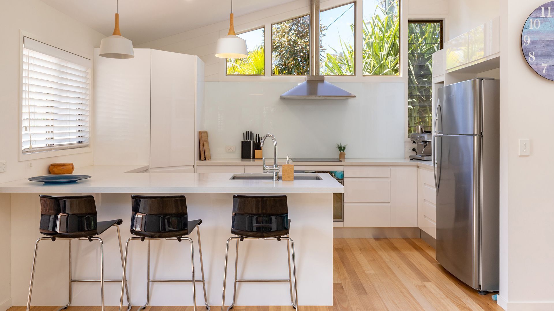 A modern kitchen with a central island, bar stools, a stainless steel refrigerator, and large windows with a view of greenery.