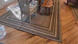 Uma área de jantar ampla com mesa e cadeiras de madeira, um tapete grande e vista para a cozinha e área externa.