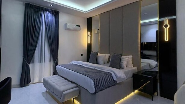 A modern bedroom with a large bed, bedside tables, a bench, a mirror, and a ceiling light fixture. - DELIZ HOMES AND HOTEL (Lekki)