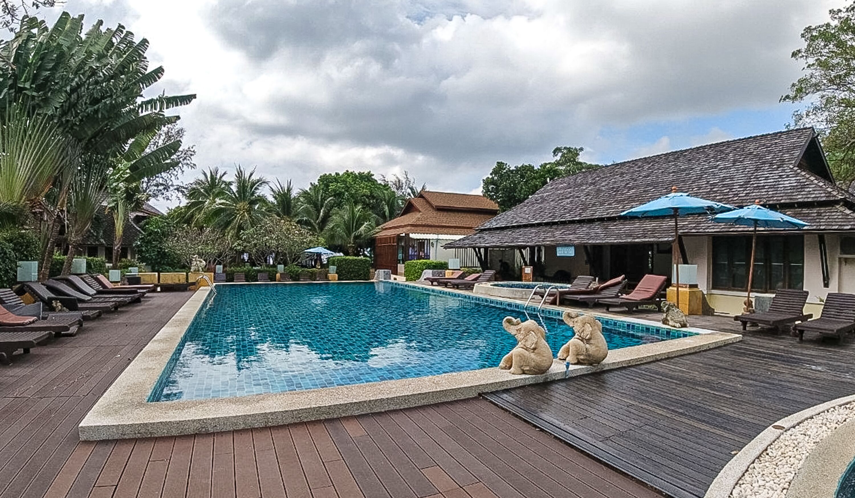 A swimming pool with lounge chairs and umbrellas, surrounded by tropical vegetation and buildings.