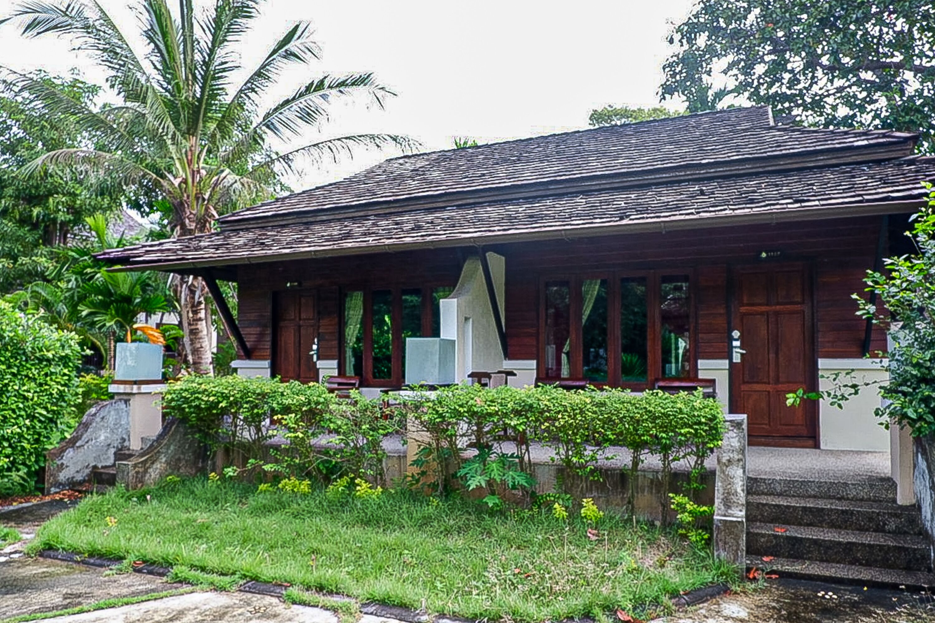 A traditional wooden building with a sloped roof, surrounded by lush greenery and palm trees.