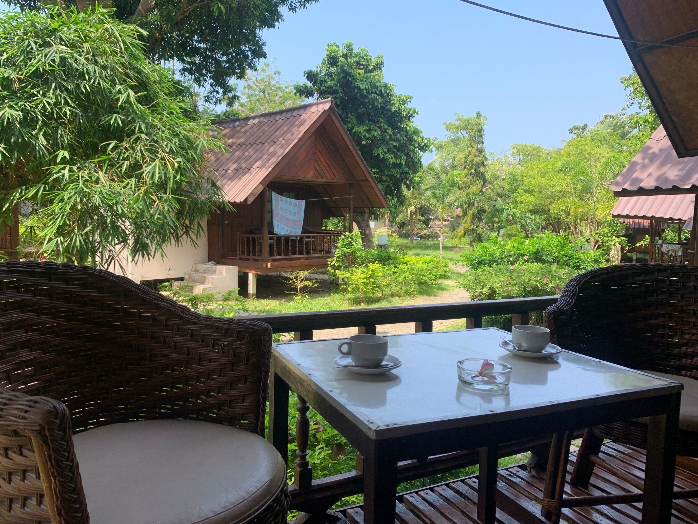 A wooden deck with wicker chairs and a table, overlooking a lush garden and a small wooden house with a balcony.