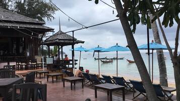 A wooden deck with tables and chairs, a gazebo, and blue umbrellas overlooking a calm sea with boats.