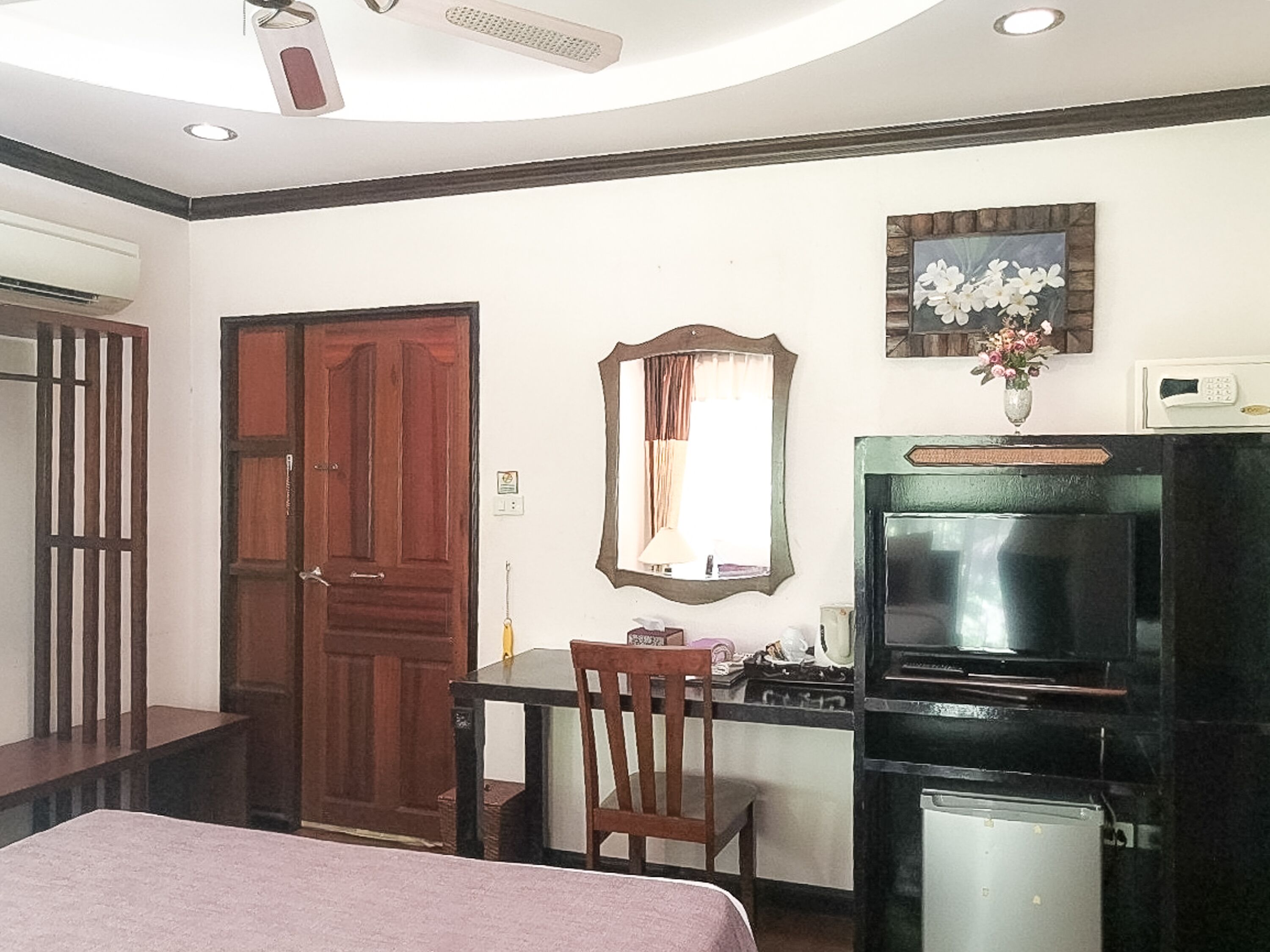 Habitación de hotel con cama, escritorio con silla, televisor, refrigerador, puerta de madera y espejo en la pared.