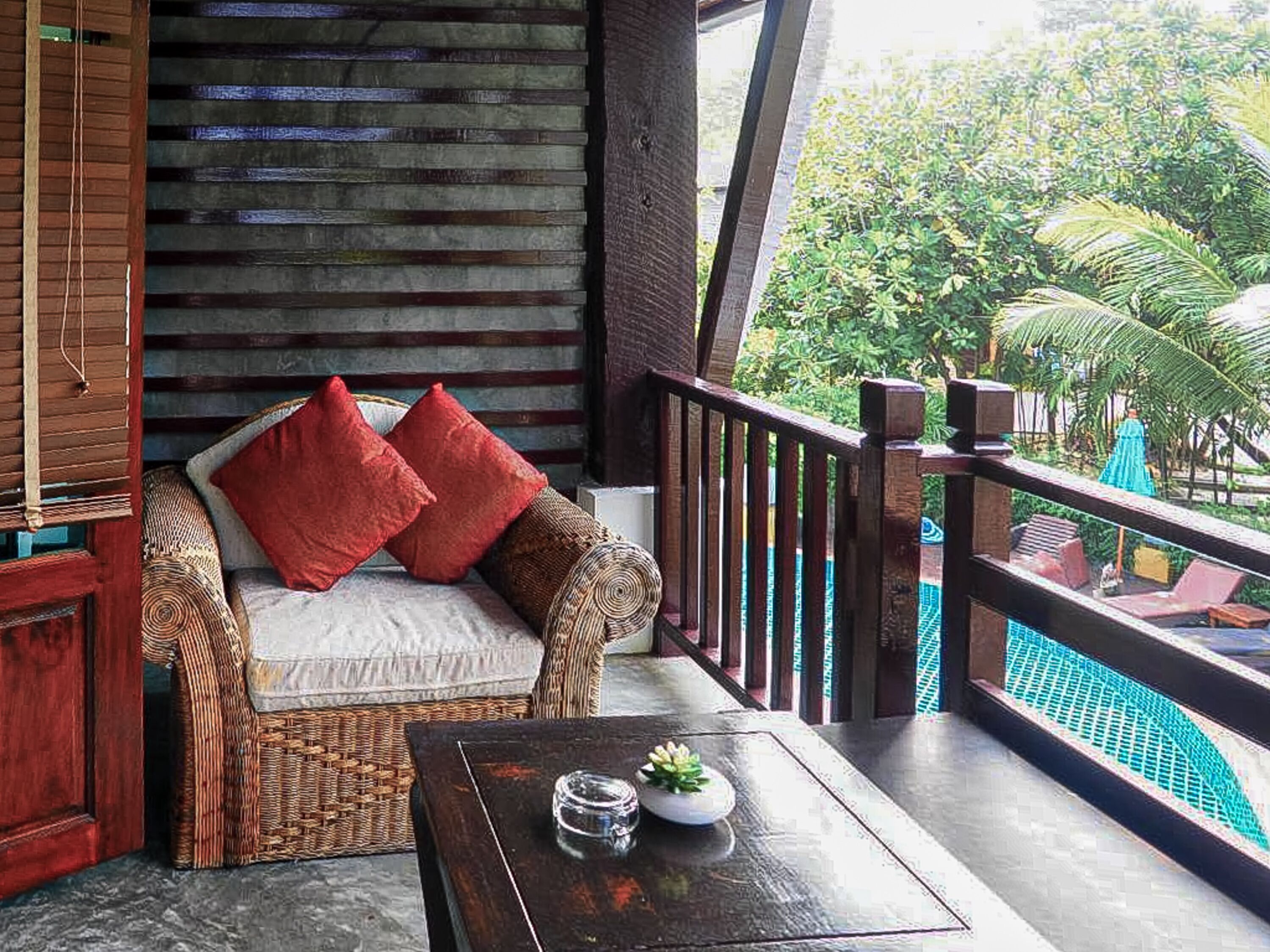 Ein Balkon mit einer Rattan-Couch, roten Kissen, einem kleinen Tisch mit einem Glas und einer Pflanze, sowie Blick auf einen Swimmingpool und Grünflächen.