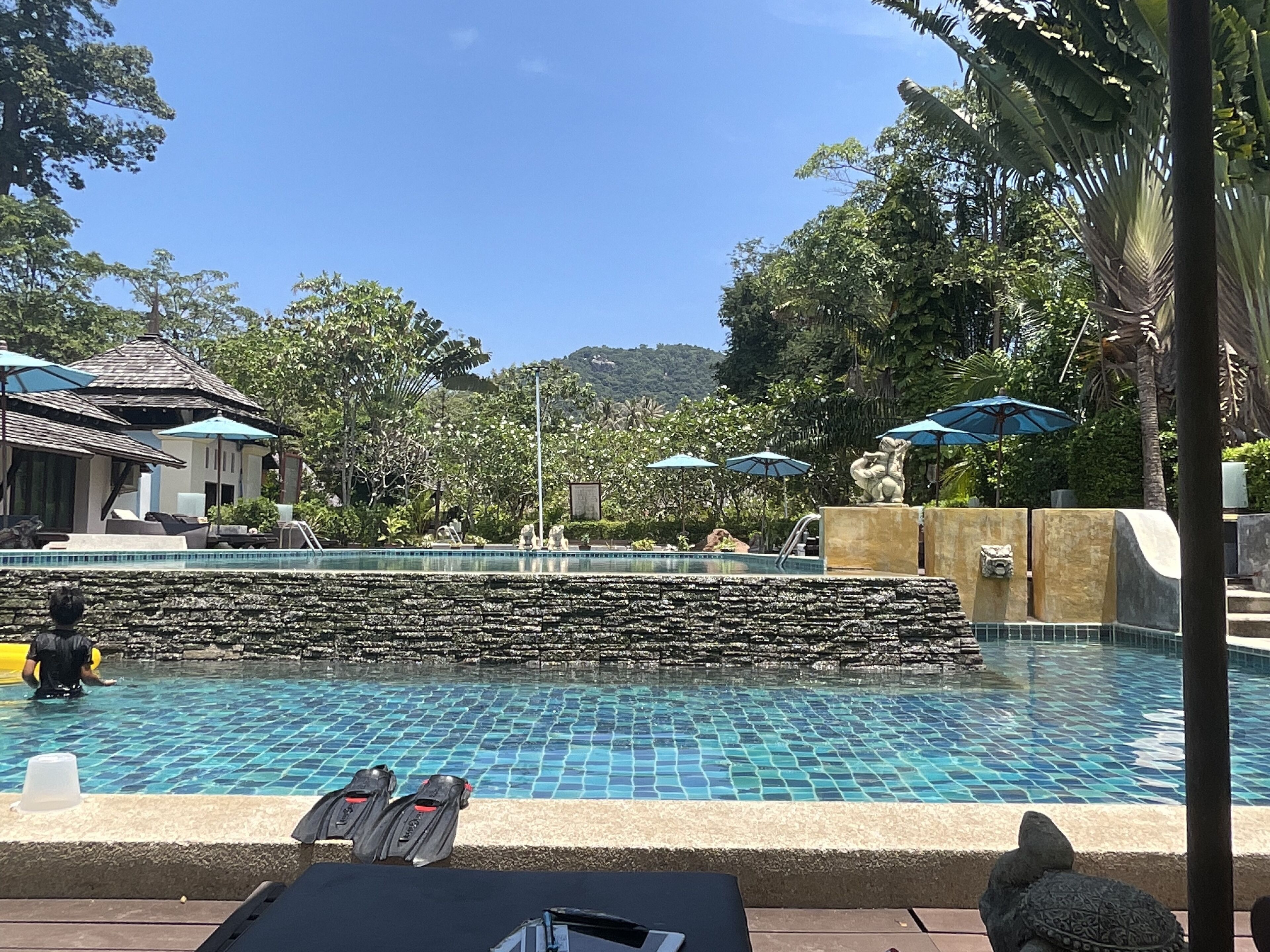 Una piscina con suelo de baldosas, rodeada de sillones de descanso y sombrillas, con un muro de piedra y plantas tropicales al fondo.