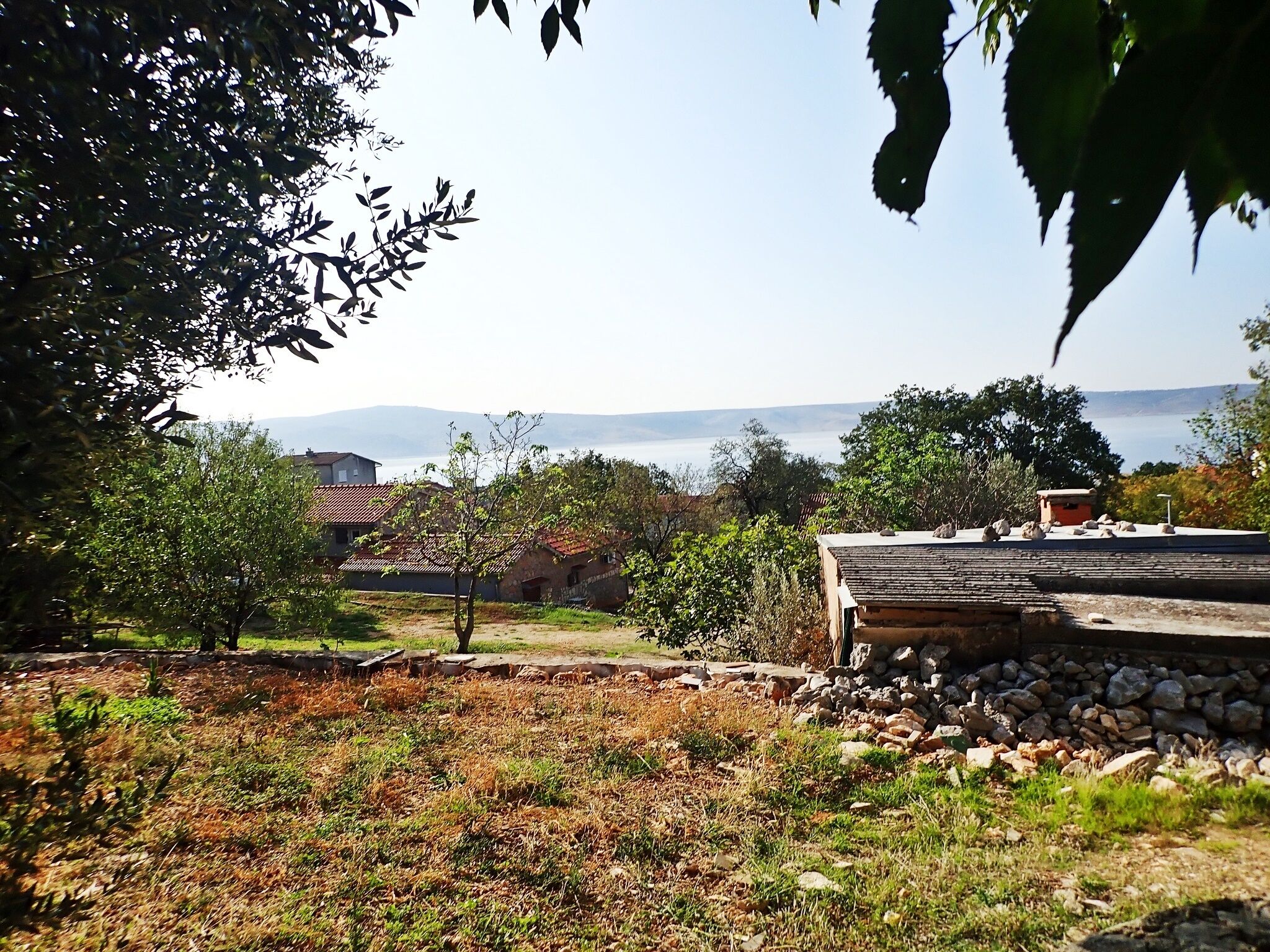 A rural landscape with a stone wall, a small building, and a view of the sea and mountains in the distance.