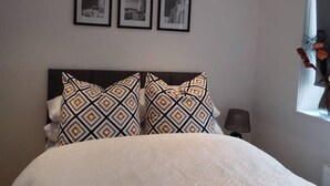 Een slaapkamer met een bed, twee kussens met een patroon, een nachtkastje, een lamp, en drie ingelijste foto’s aan de muur.